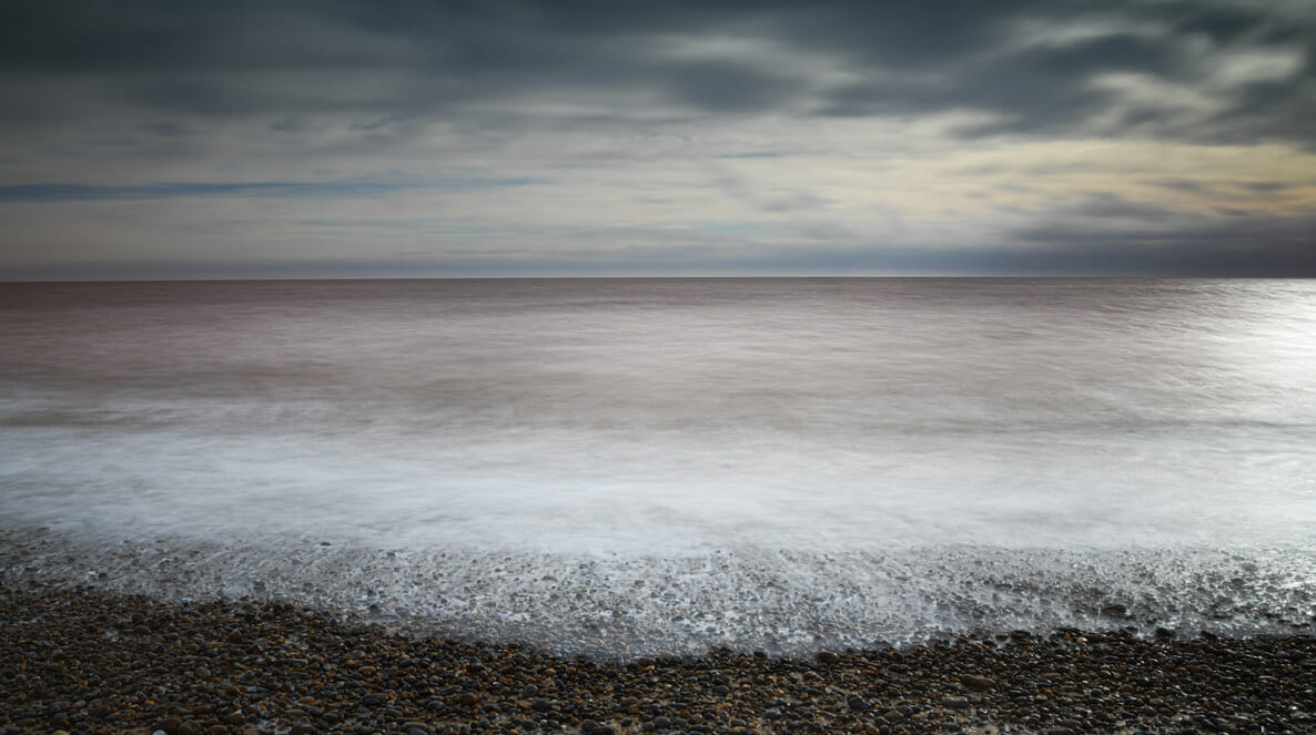 A colour photograph of a foamy sea breaking on a pebbled beach with sky and clouds above, by Susi Petherick